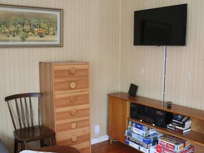 Une pièce avec une commode en bois, un téléviseur fixé au mur et une bibliothèque remplie de livres et de jeux de réflexion.