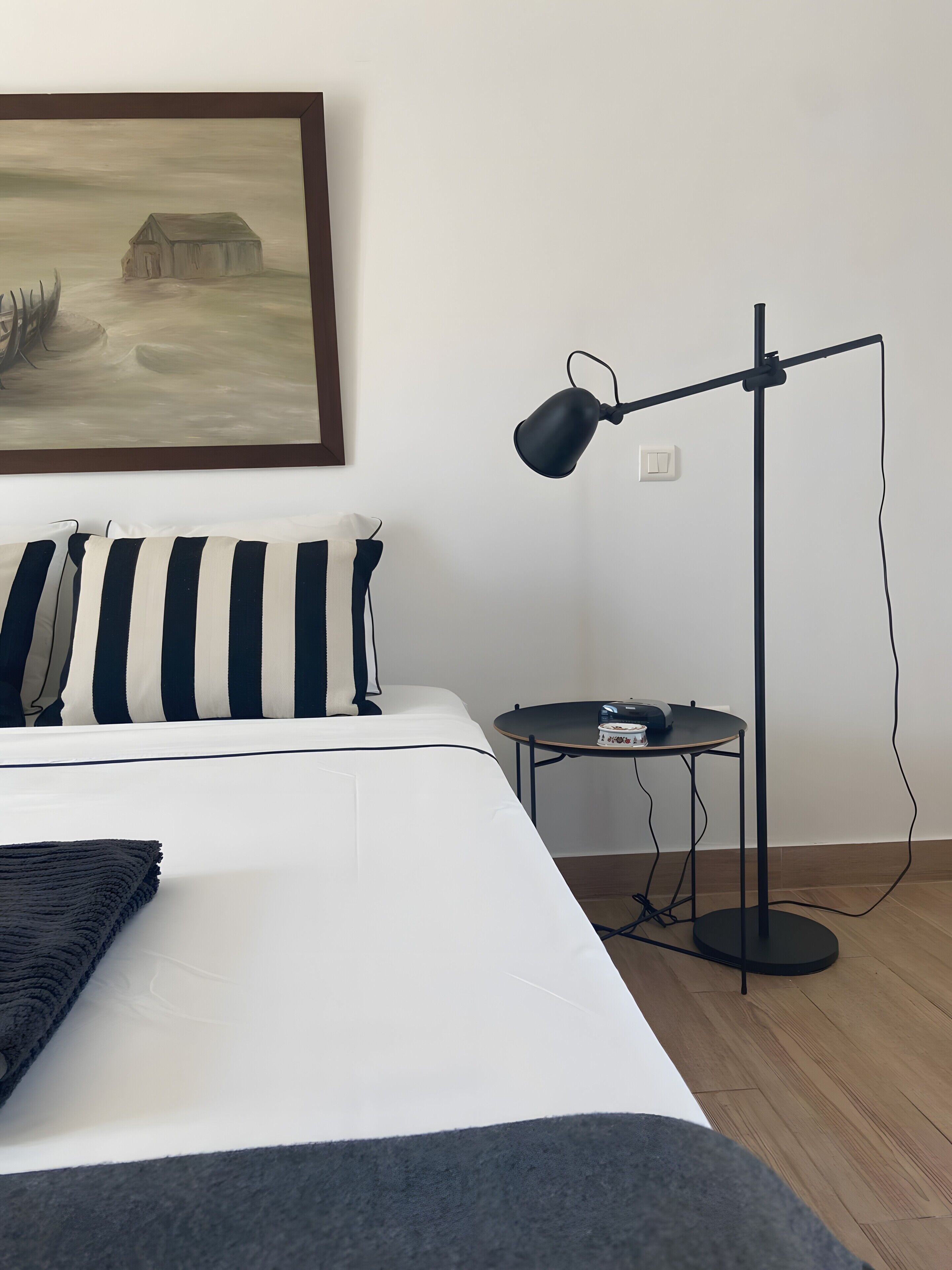 A neatly made bed with striped pillows, a standing lamp, a small table with a device, and a painting on the wall.