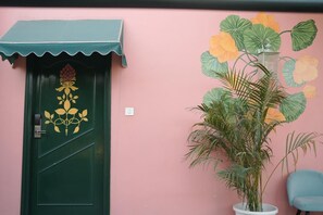 A green door with a decorative pattern, a potted plant with large leaves, and a blue chair against a pink wall. - Artistik Abode By Stay Banaras (Varanasi)