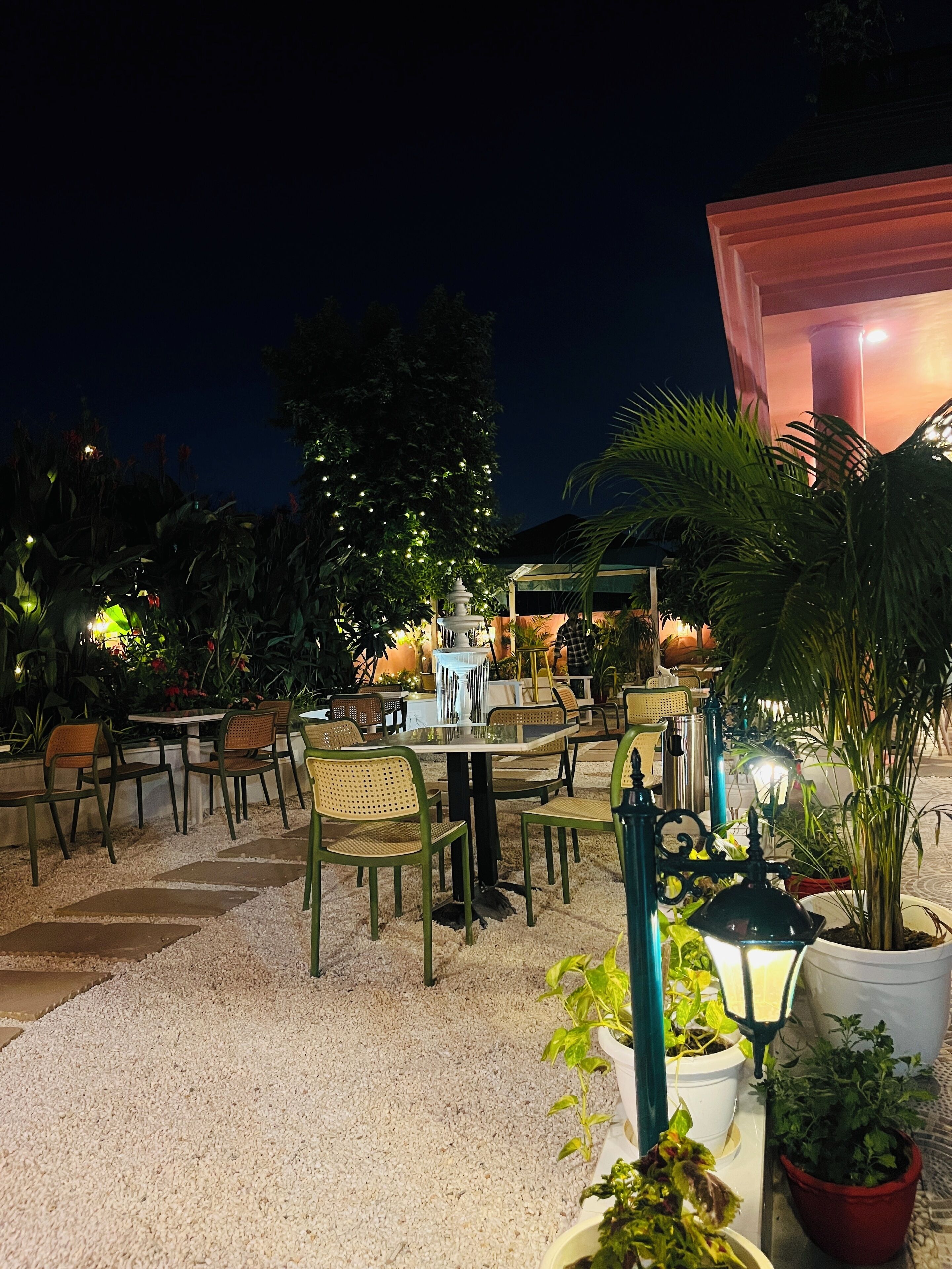 Un espace extérieur aménagé avec des tables et des chaises, entouré de plantes en pot et agrémenté d’une fontaine décorative.