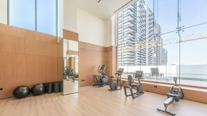 A gym with exercise equipment and a large mirror, a wooden floor, and a view of a high-rise building. - JVC 1 bed Sleeps 2 With Private Balcony (Dubai)