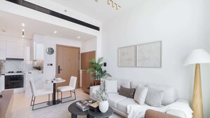 A modern living room with a sofa, coffee table, and chairs. There is a dining area with a table and chairs. The kitchen is visible in the background with white cabinets and a stove. - JVC 1 bed Sleeps 2 With Private Balcony (Dubai)