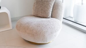 Une chaise moderne, ronde, de couleur beige, recouverte de tissu, avec un piétement doré, placée dans une pièce lumineuse dotée d’une grande fenêtre et d’une petite table d’appoint blanche.