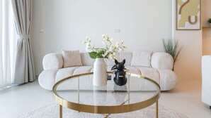 Un salon moderne avec une table basse ronde en verre, un vase blanc orné de fleurs, une statuette de chat noir et un canapé beige.