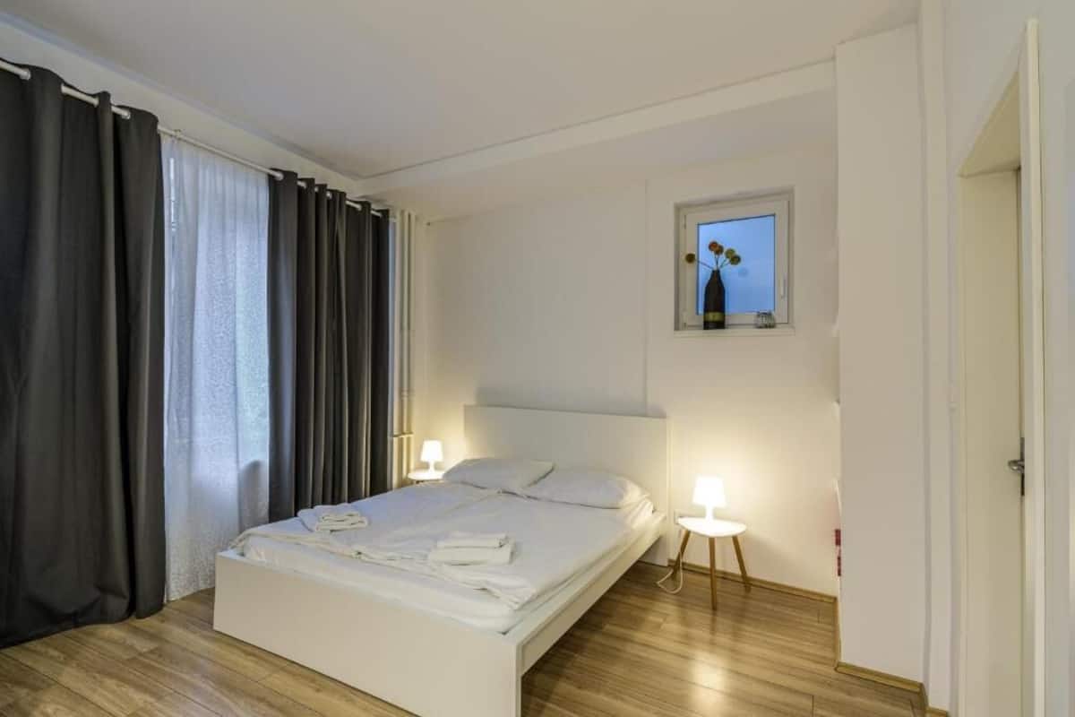 Un letto rifatto con cura in una stanza con pavimento in legno, una piccola finestra con un vaso e due abat-jour.