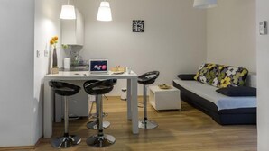 Ein kompakter Wohnraum mit Küchenzeile, einer Essecke mit zwei Barhocker und einem Sofa mit floralem Kissen sowie einem kleinen Beistelltisch.
