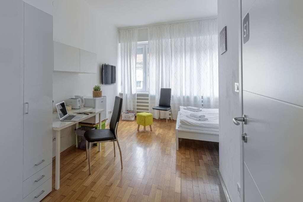 Een moderne, goed verlichte kamer met een houten vloer, een bureau met een laptop, een stoel, een bed en een raam met dunne gordijnen.