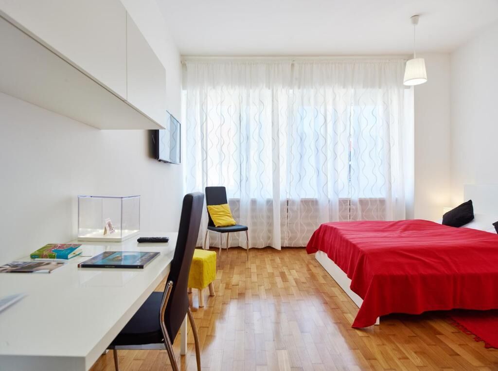 Una habitación moderna con un escritorio blanco, una silla, una cama con colcha roja y una ventana con cortinas blancas.