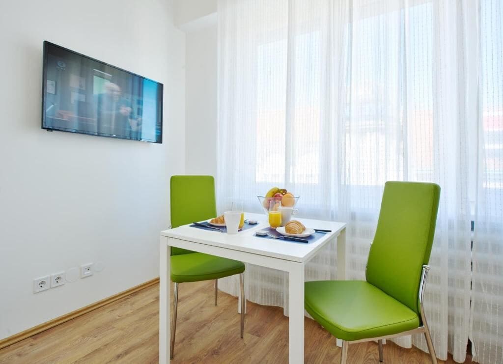 Un pequeño comedor con una mesa blanca, dos sillas verdes y una disposición para el desayuno que incluye un croissant, café y jugo de naranja.