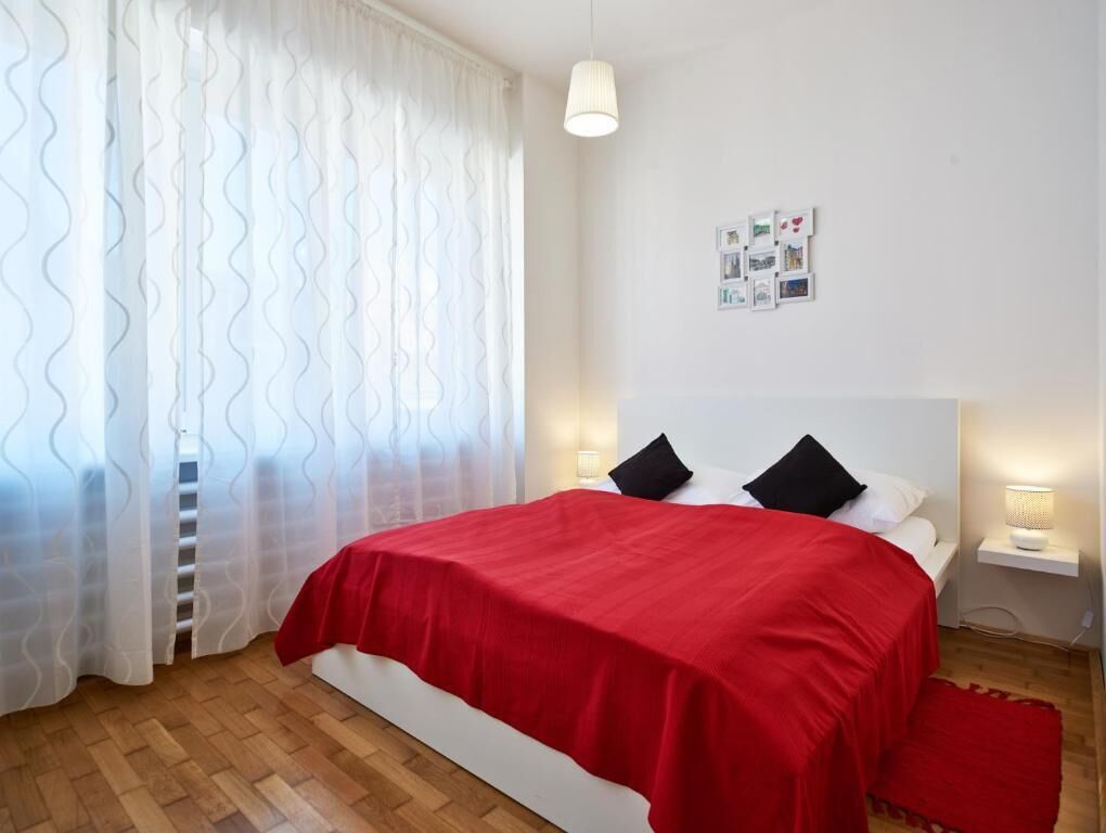 Un dormitorio con una cama, cortinas blancas, un cuadro en la pared y una lámpara de noche.