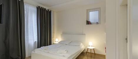A modern bedroom with a large bed, two bedside lamps, a window with curtains, and a small bedside table with books.