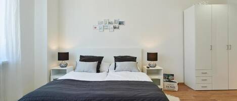 A modern bedroom with a large bed, two bedside tables with lamps, a wardrobe, and a wall with framed pictures.