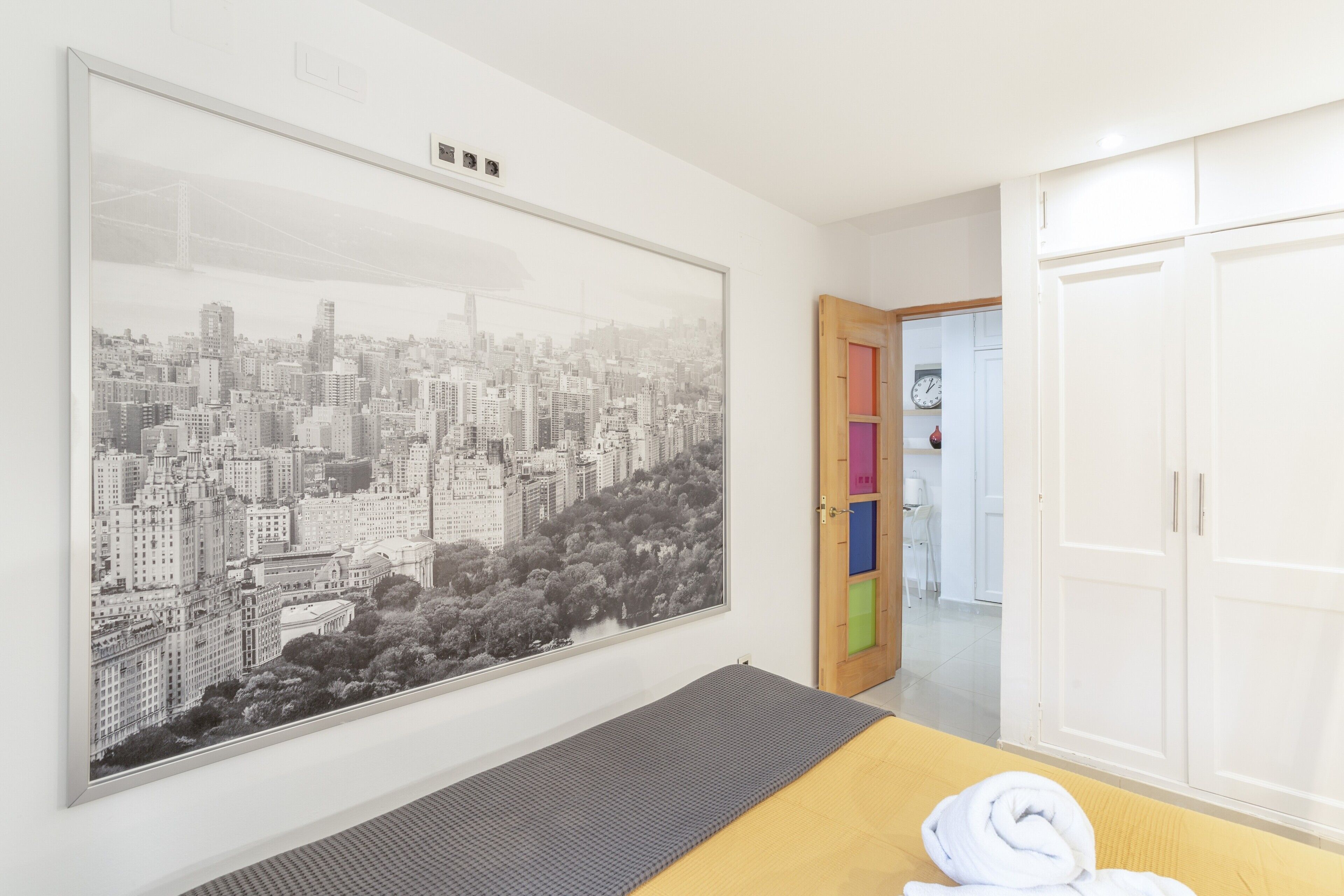 Una habitación con una gran obra de arte de un paisaje urbano, una cama con un colchón gris, una puerta de madera con paneles de colores y armarios blancos.