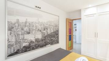 Una habitación con una gran obra de arte de un paisaje urbano, una cama con un colchón gris, una puerta de madera con paneles de colores y armarios blancos.