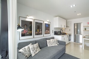 Un salon moderne avec un canapé gris, un coin repas avec une table blanche et une cuisine avec des meubles blancs et un réfrigérateur en acier inoxydable.