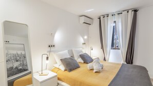 Hotelkamer met een bed, nachtkastje, lamp, airconditioning en een raam met gordijnen, met uitzicht op de stad.