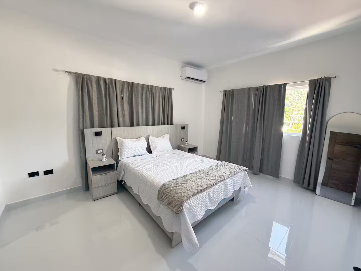 Ein modernes Schlafzimmer mit einem großen Bett, Nachttischen, einer Klimaanlage und Blick ins Freie durch ein Fenster mit Vorhängen.