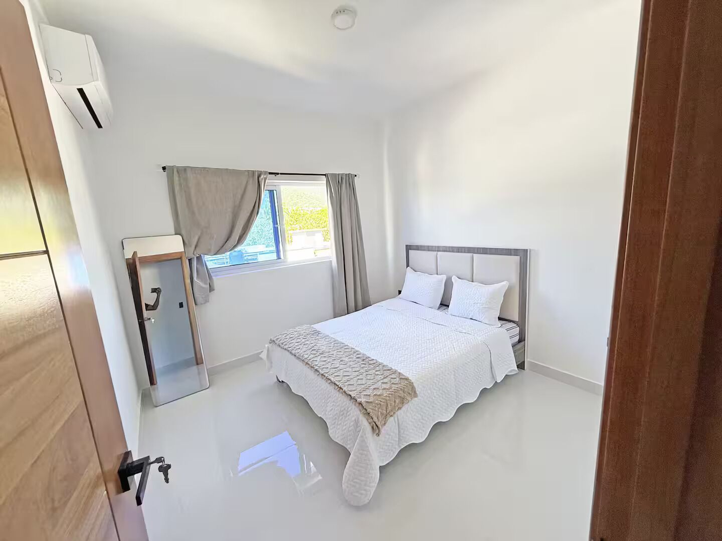 A bedroom with a bed, a window with curtains, a bedside table, and a wall-mounted air conditioner.