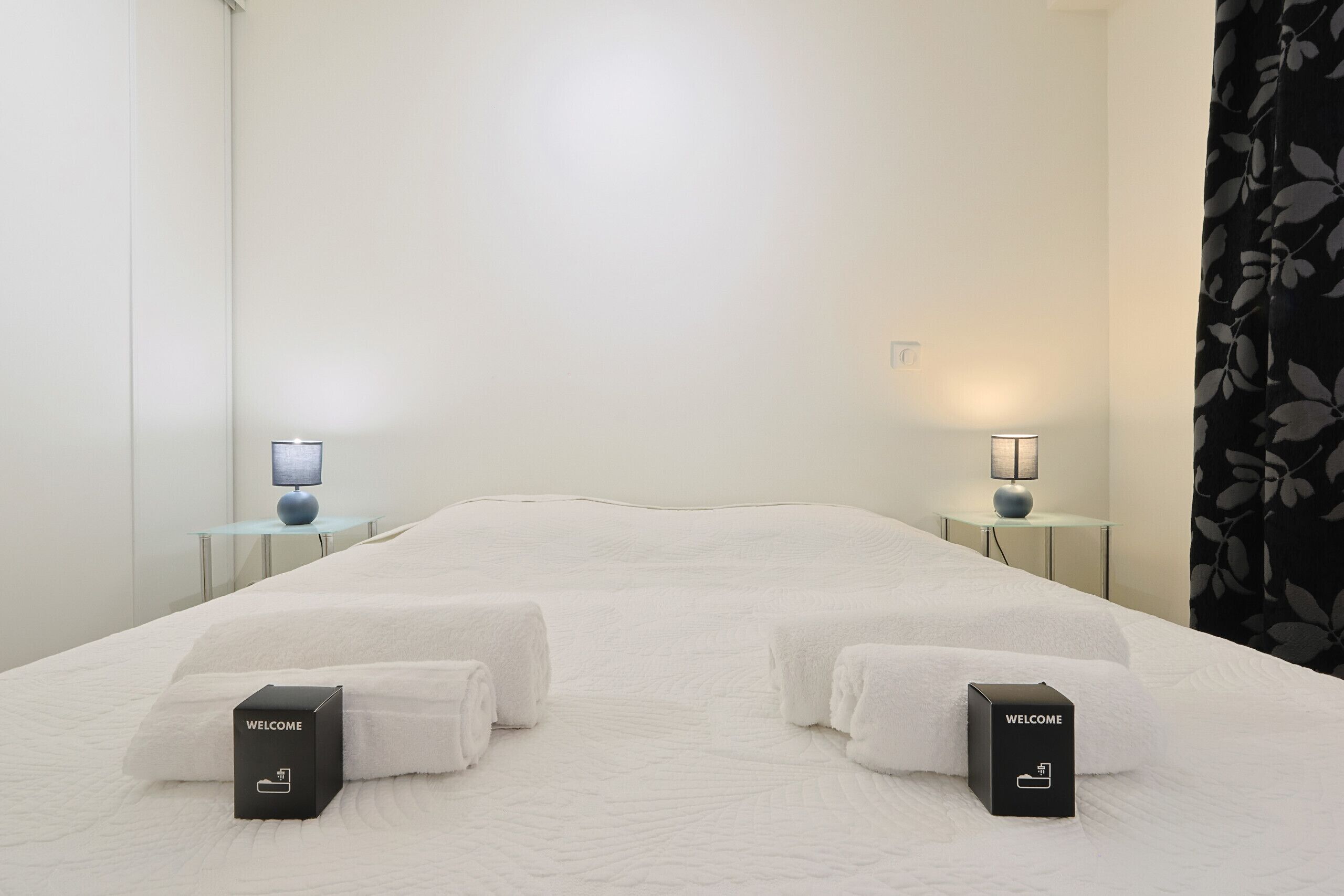 Una habitación de hotel con una cama grande, dos mesitas de noche con lámparas y dos cajas de regalo de bienvenida.
