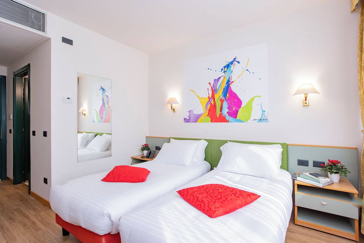 Ein Hotelzimmer mit zwei Betten, roten Kissen, einem grünen Kopfteil, einem Nachttisch mit Lampe und einem farbenfrohen abstrakten Gemälde an der Wand.