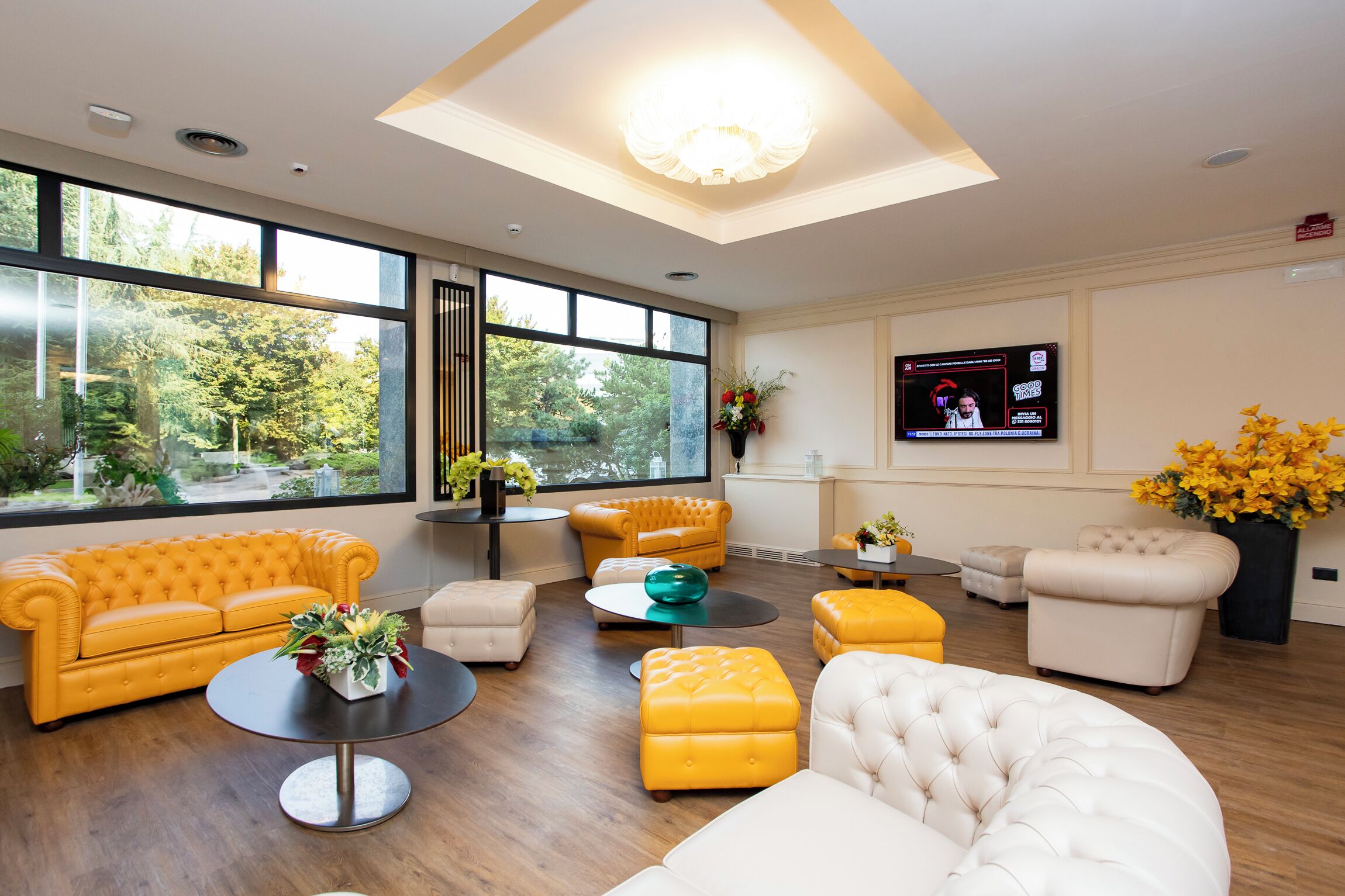 A modern living room with large windows, a flat-screen TV, and a variety of seating options including a yellow sofa, a white armchair, and a round coffee table.