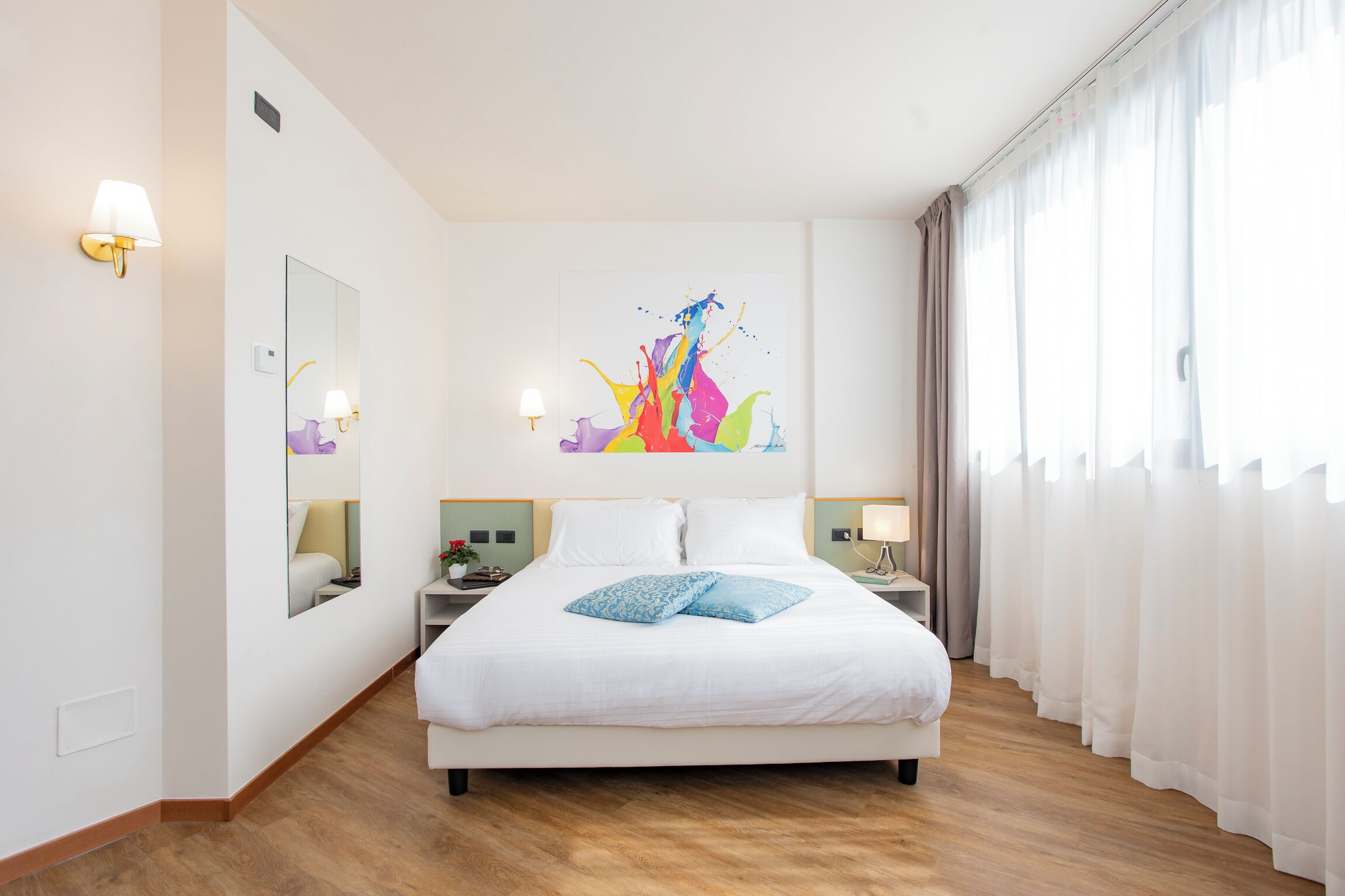 Una camera d'albergo con un letto grande, uno specchio, un comodino, una lampada e un quadro astratto dai colori vivaci appeso alla parete.