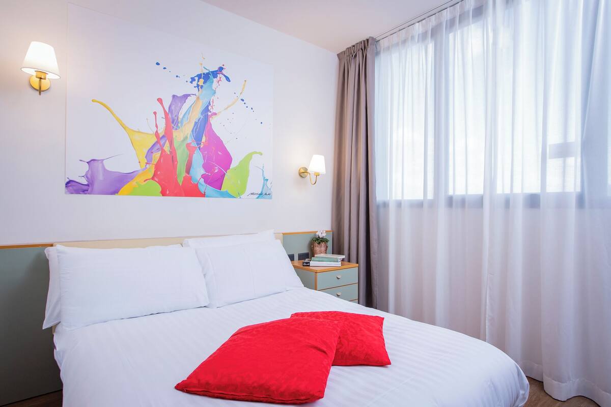 Ein ordentlich bezogenes Bett mit weißen und roten Kissen, ein farbenfrohes abstraktes Gemälde an der Wand und ein Nachttisch mit Lampe und Pflanze.