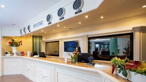 Un espace d’accueil d’hôtel avec un comptoir de réception blanc, une horloge au-dessus et une pancarte de bienvenue.