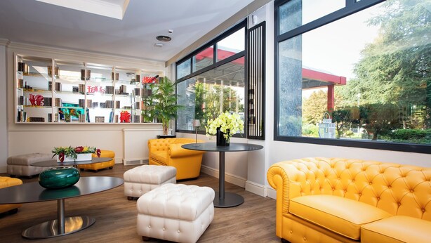 Un salon moderne avec un canapé jaune, un pouf blanc et une table ronde ornée d’un vase de fleurs. On y trouve une bibliothèque remplie de divers objets et une grande fenêtre donnant sur l’extérieur.