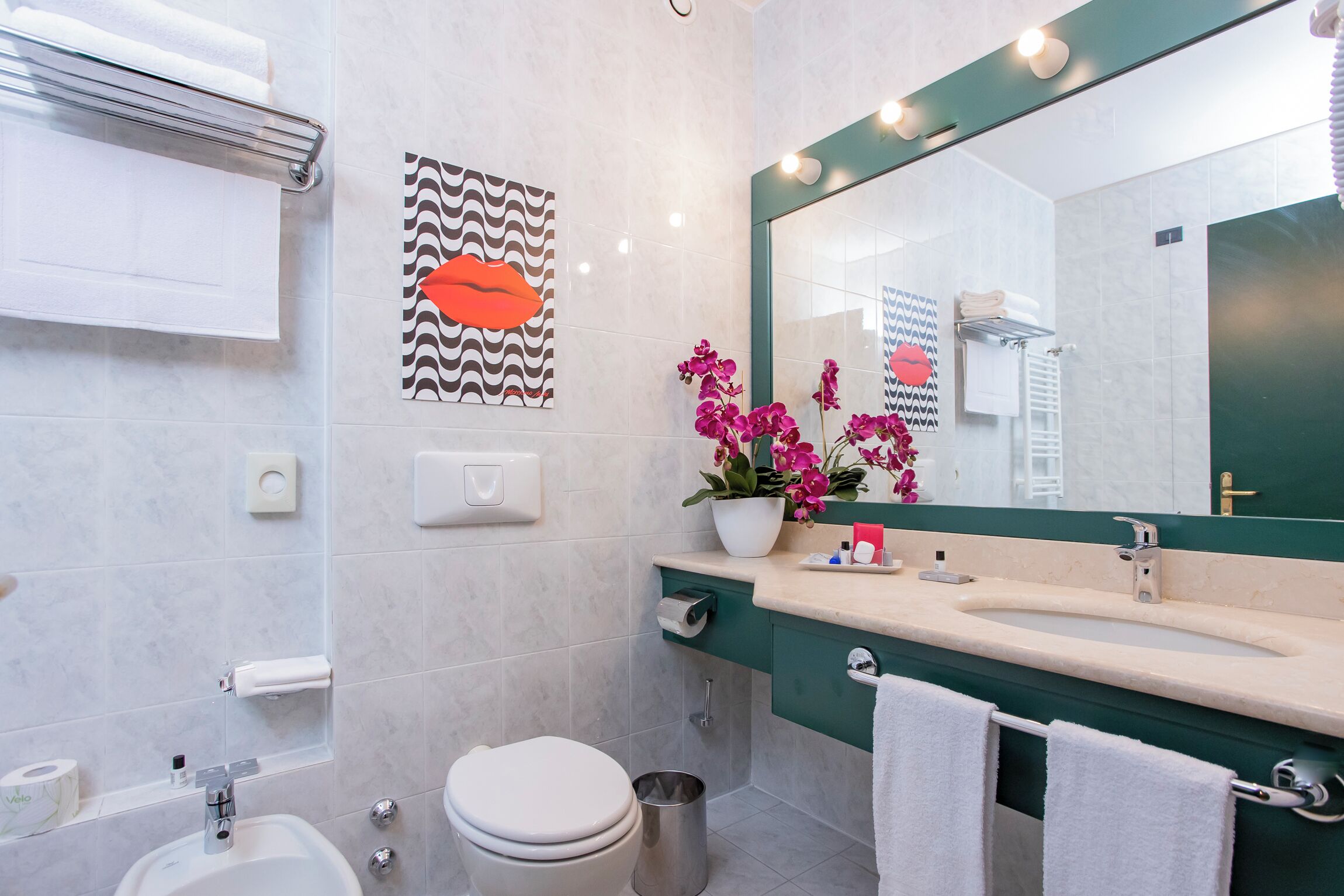 Ein modernes Badezimmer mit großem Spiegel, weißen Fliesen, einem Waschbecken, einer Toilette und einem Orchideenstrauch.