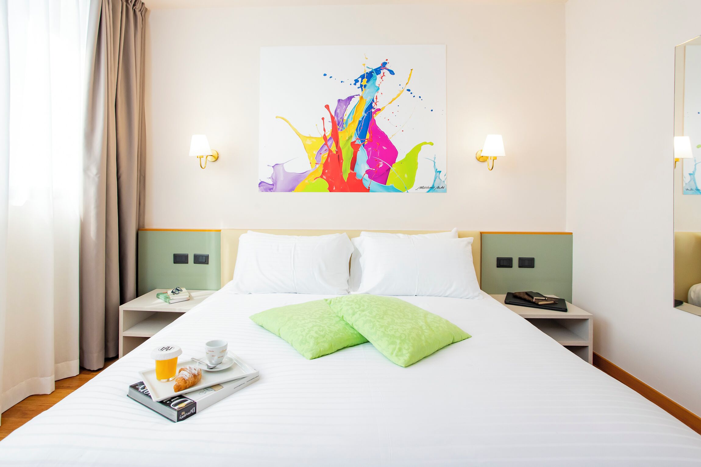 A hotel room with a bed, green pillows, a tray with a drink and pastry, and a colorful abstract painting on the wall.