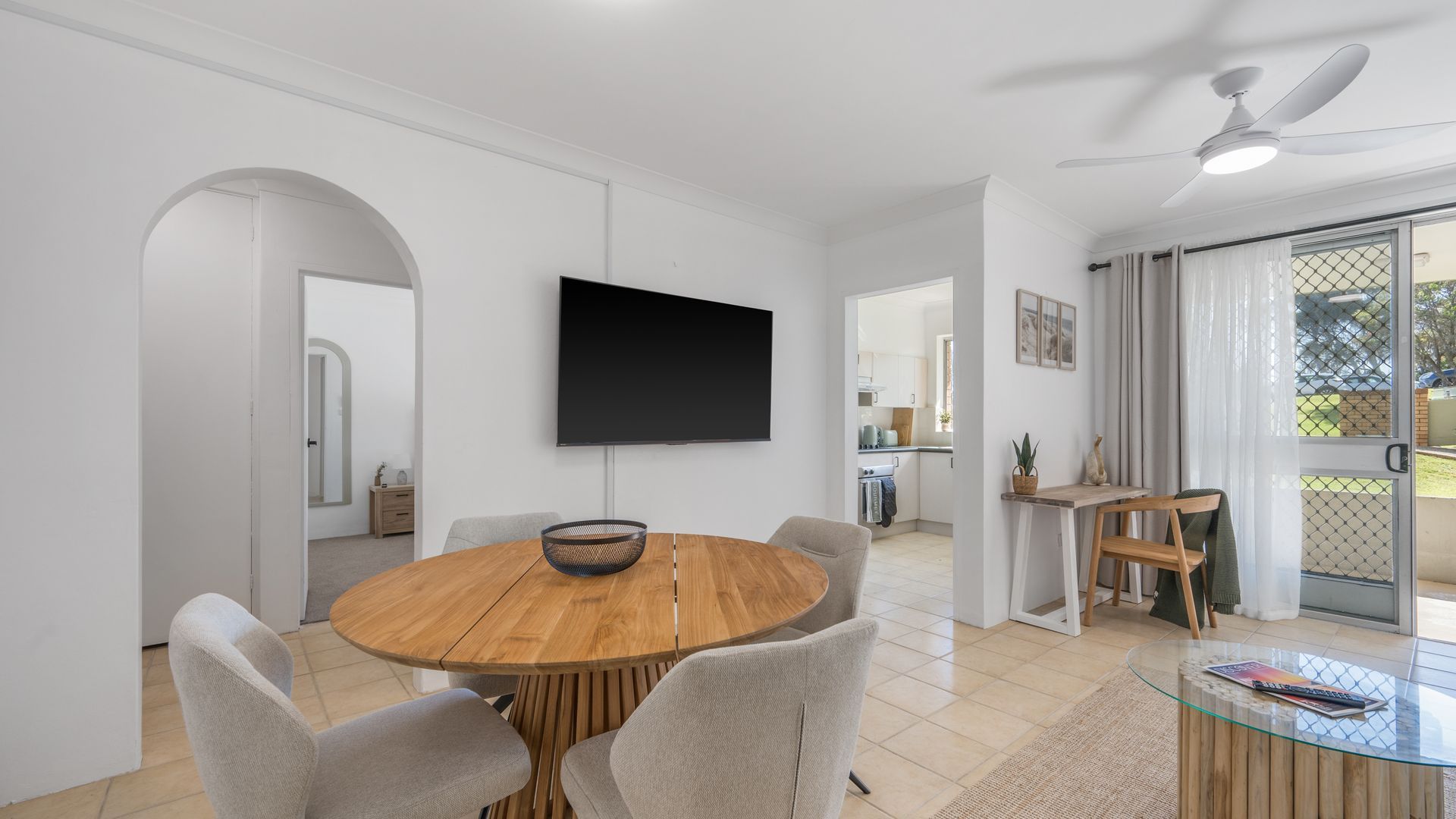A modern dining area with a round wooden table, grey upholstered chairs, a flat-screen TV mounted on the wall, and a ceiling fan.