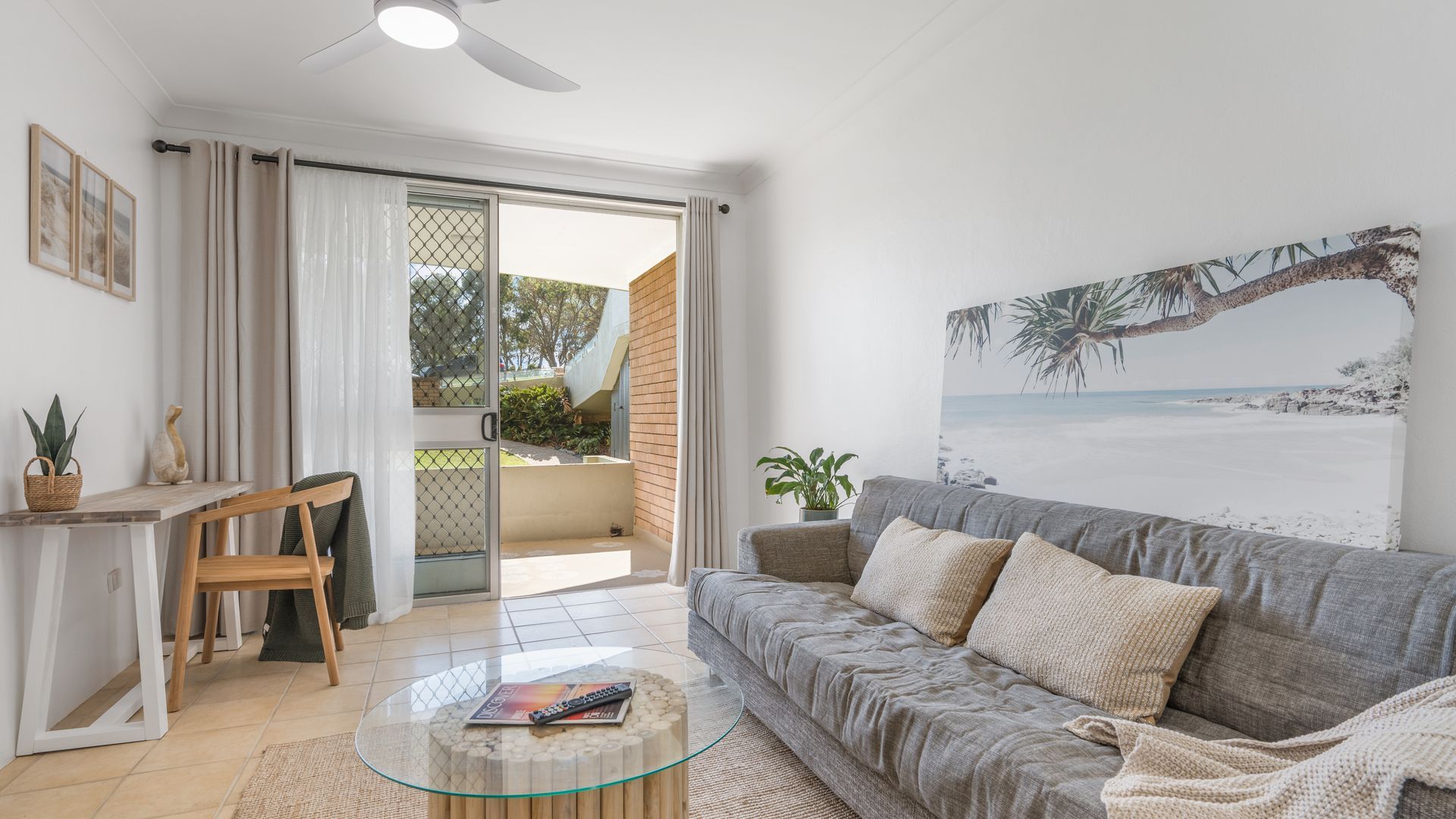 A living room with a grey sofa, a glass coffee table, and a painting of a beach scene.