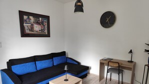 Uma sala de estar moderna com um sofá preto e azul, uma mesa de centro de madeira, uma escrivaninha com cadeira, um relógio e uma luminária pendente.
