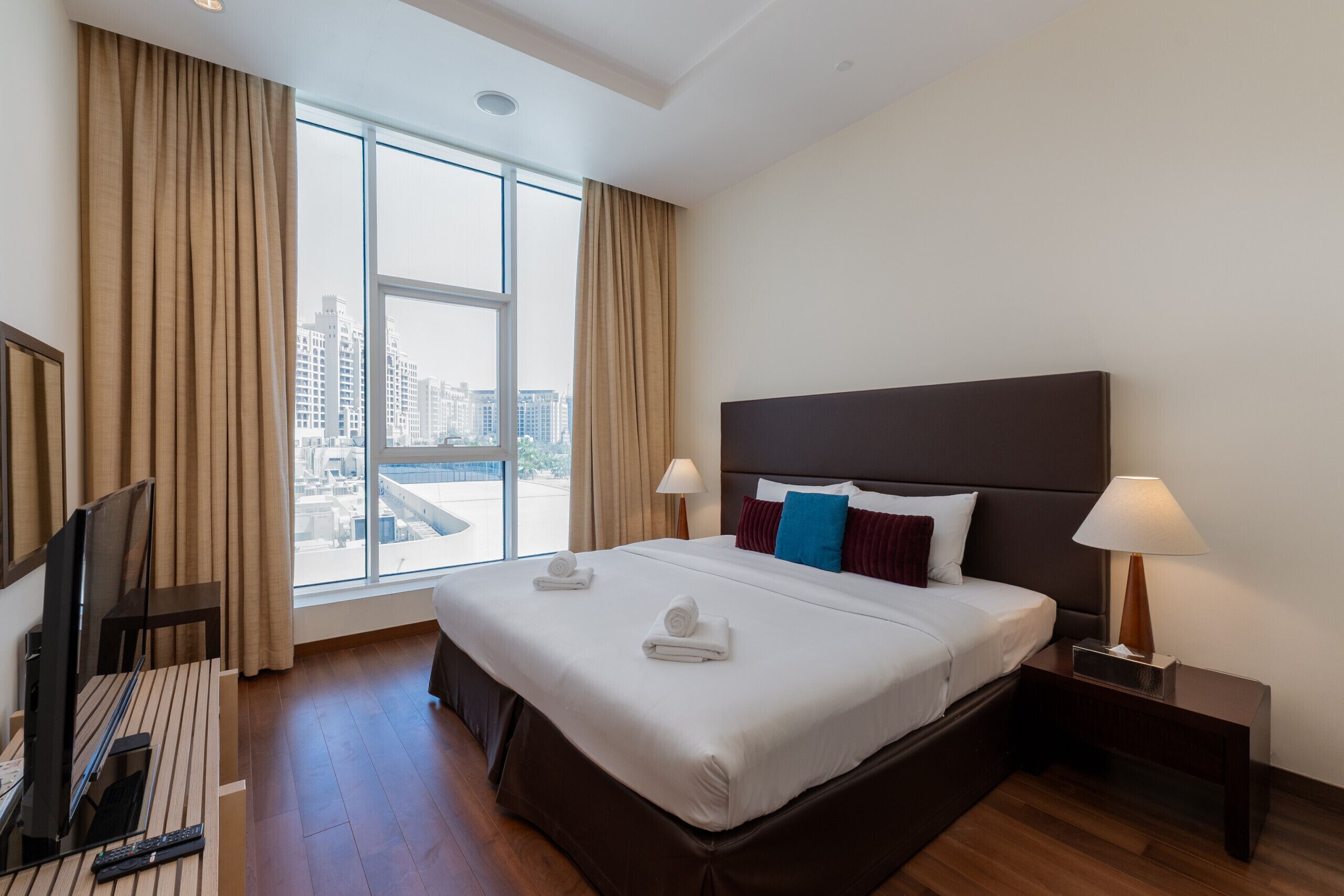 Quarto de hotel com uma cama grande, duas luminárias de cabeceira, televisão de tela plana e vista da cidade por grandes janelas.
