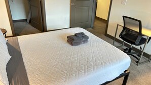 A neatly made bed with a white quilt and folded gray towels, a black office chair, and a closed door in a hotel room. - Boulevard Stays Contemporary 1BR Apartment, Free Parking (Louisville)