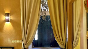 A hallway with a patterned floor leading to a dark door, beige curtains, and a wall-mounted light fixture.