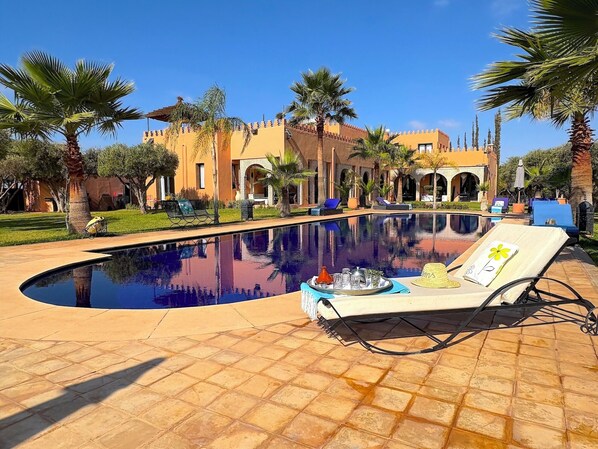 A pool area with lounge chairs, palm trees, and a building with arches in the background. - Palais Khemia (Marrakech)