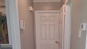 Un couloir étroit, avec une porte blanche au fond, un tapis blanc sur le sol et un interrupteur sur le mur.