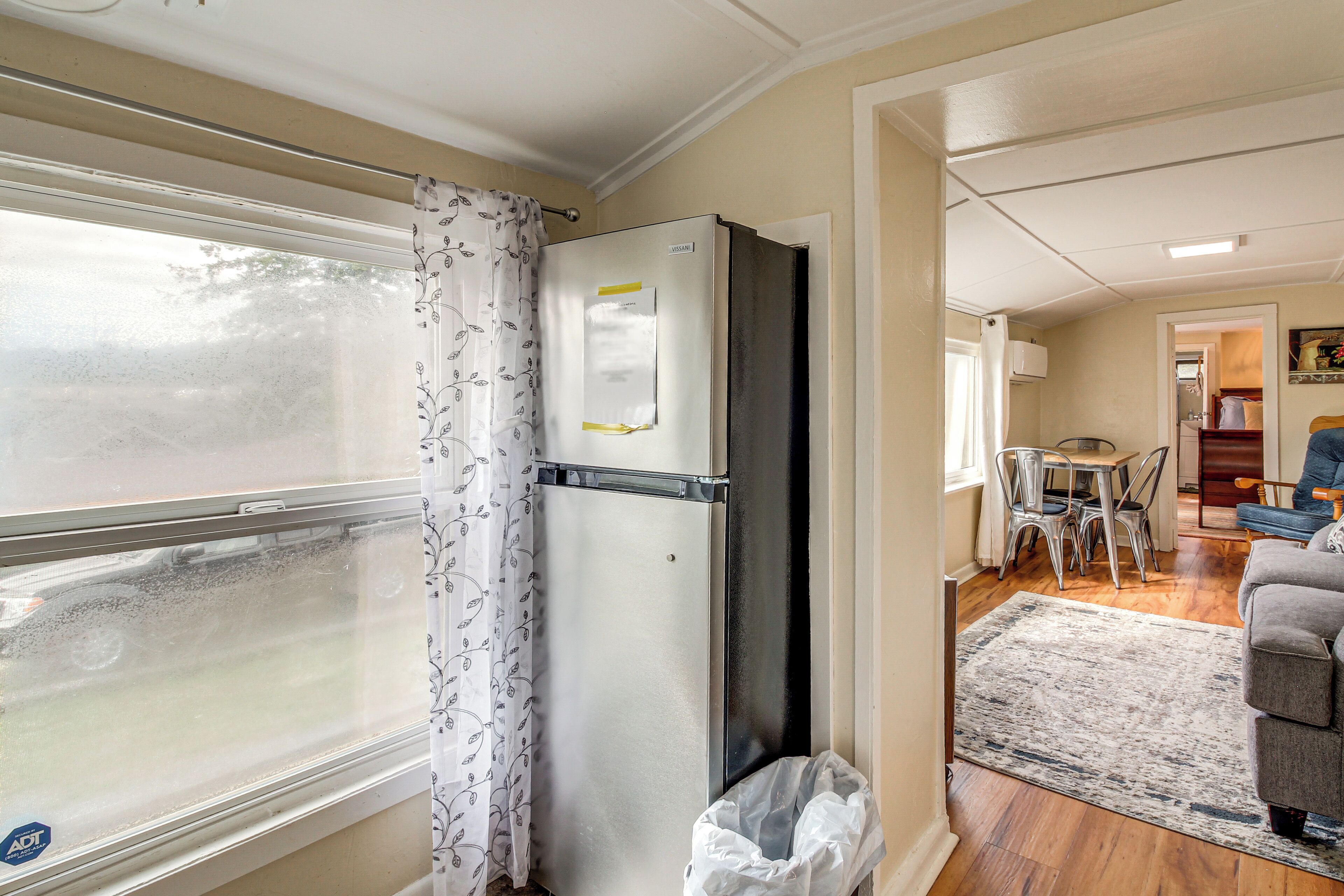 A compact kitchen with a refrigerator, a window with a curtain, a dining area with a table and chairs, and a living room with a sofa.