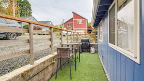 A patio area with a wooden deck, two chairs, a grill, and a blue building with a window. - Quaint Coos Bay Escape, 2 Mi to Downtown (Coos Bay)