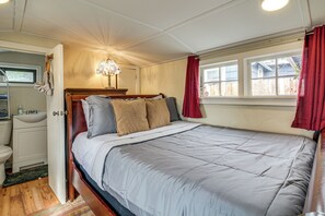 A bedroom with a bed, a nightstand, a lamp, a window with curtains, and a view of the outside. - Quaint Coos Bay Escape, 2 Mi to Downtown (Coos Bay)