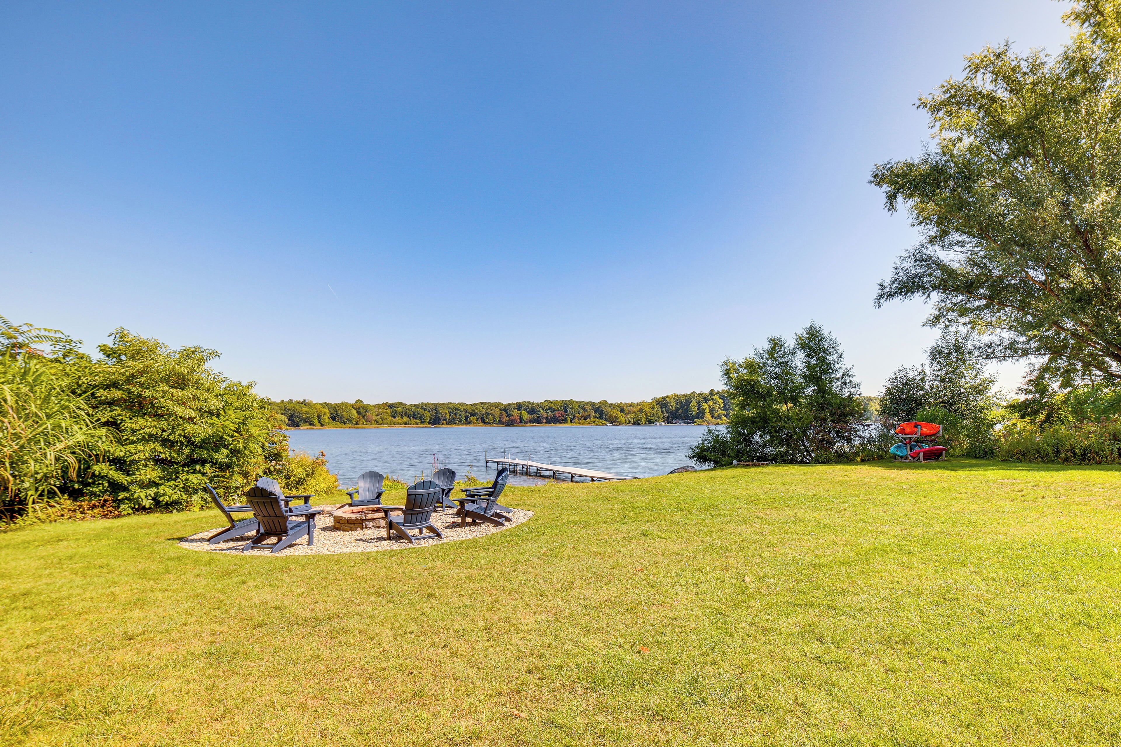A lakeside patio with Adirondack chairs and a fire pit, surrounded by lush greenery and a dock extending into the water.