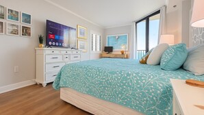 Ein Schlafzimmer mit einem Bett, einem Fernseher, einem Schminktisch und einem Fenster mit Vorhängen.