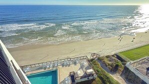 Blick auf einen Strand vom Balkon eines Hotels mit Schwimmbad und Liegestühlen.
