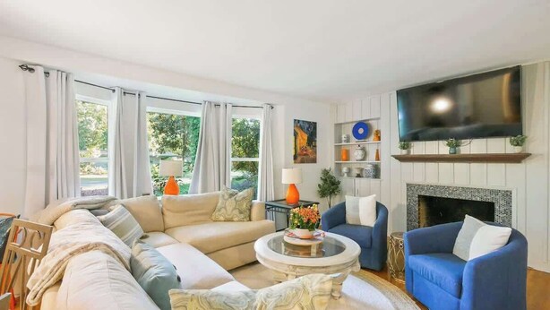 Un salón con un sofá color beige, un sillón azul, una mesa de centro redonda y un televisor de pantalla plana sobre una chimenea.