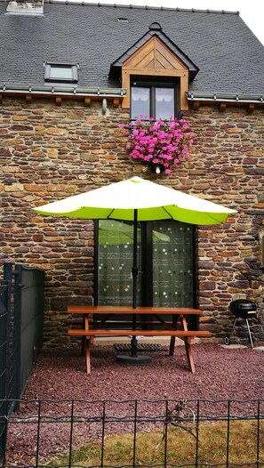 Ein Steinhaus mit einer Holzbank unter einem grünen Sonnenschirm, einer Fensterbank mit rosa Blumen und einem Grill.