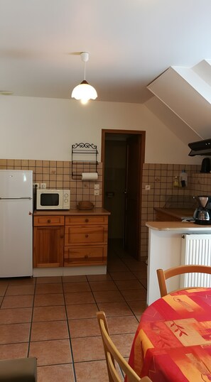 A compact kitchen with a refrigerator, microwave, wooden cabinets, a sink, and a dining area with a table and chairs. - Cottages at the Edge of Brocéliande - 4 People (Concoret)
