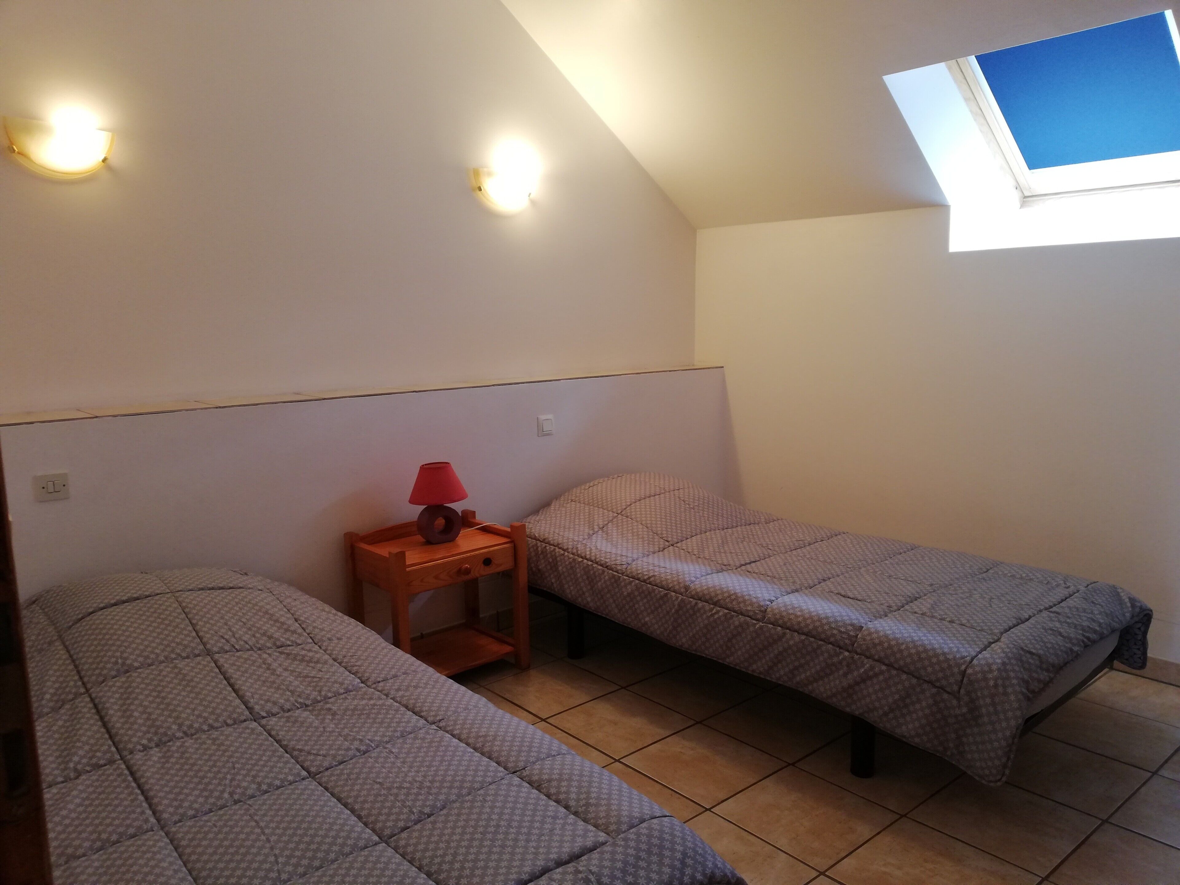 Une petite pièce avec deux lits, une table de chevet avec une lampe, une fenêtre et un luminaire au plafond.