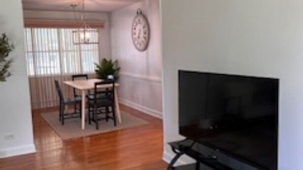A living room with a TV on a stand, wooden flooring, a dining area with a table and chairs, and a clock on the wall. - Tranquil 3-bedroom house with AC in Elk Grove Village (Elk Grove Village)